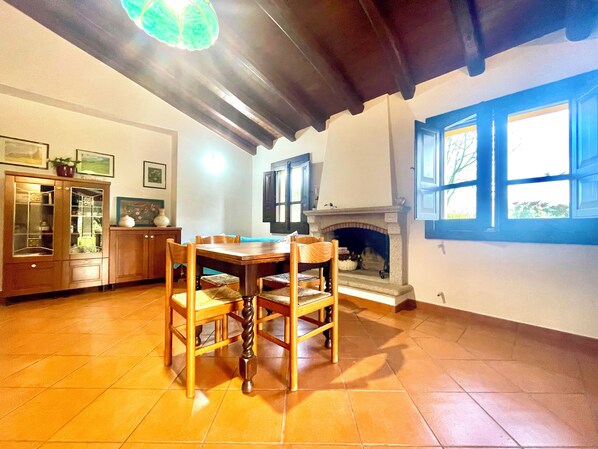 A dining area with a wooden table and chairs, a cabinet with glass doors, a fireplace, and a window with blue shutters.