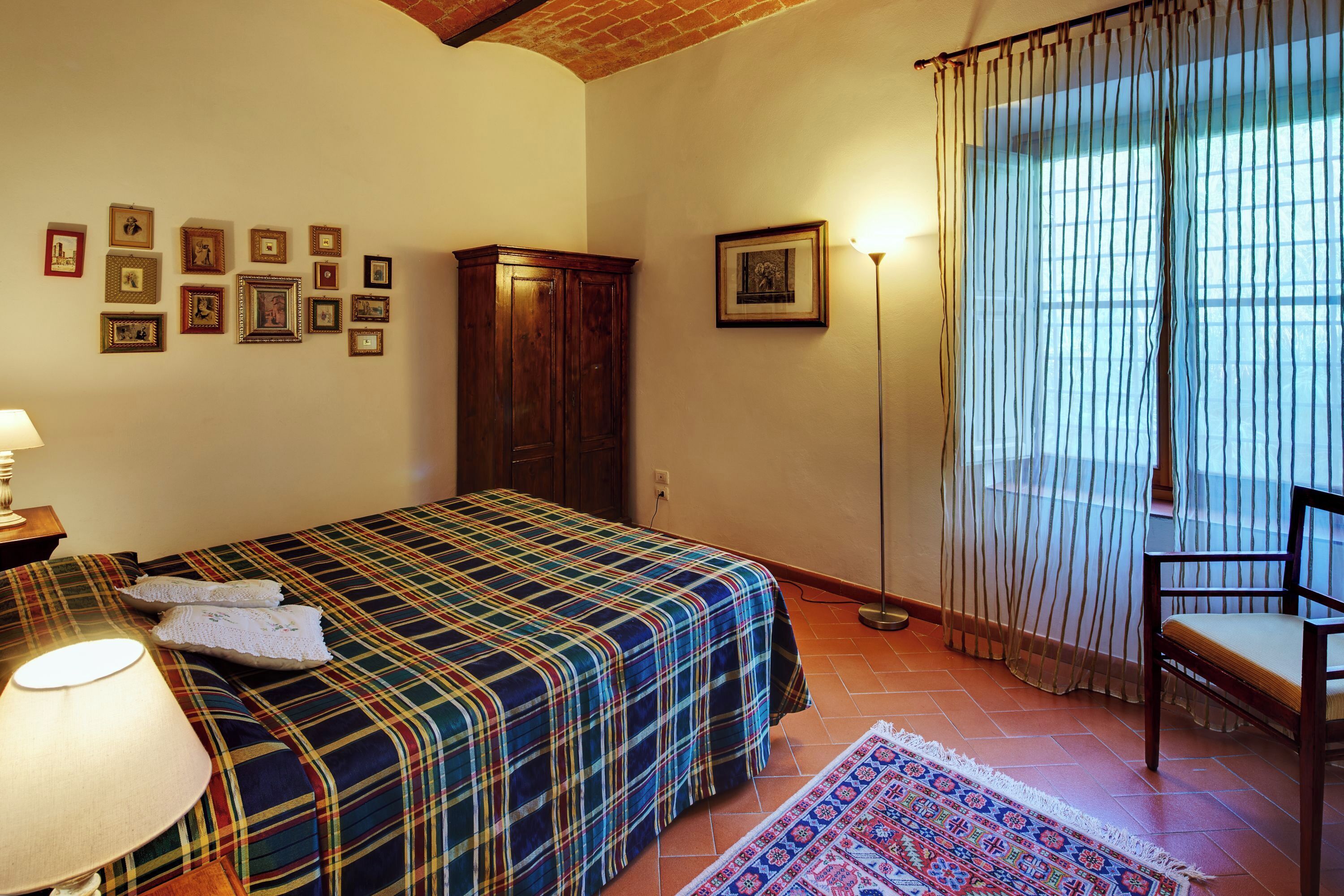 Un dormitorio con cama, armario de madera, lámpara, silla, alfombra y ventana con cortinas.