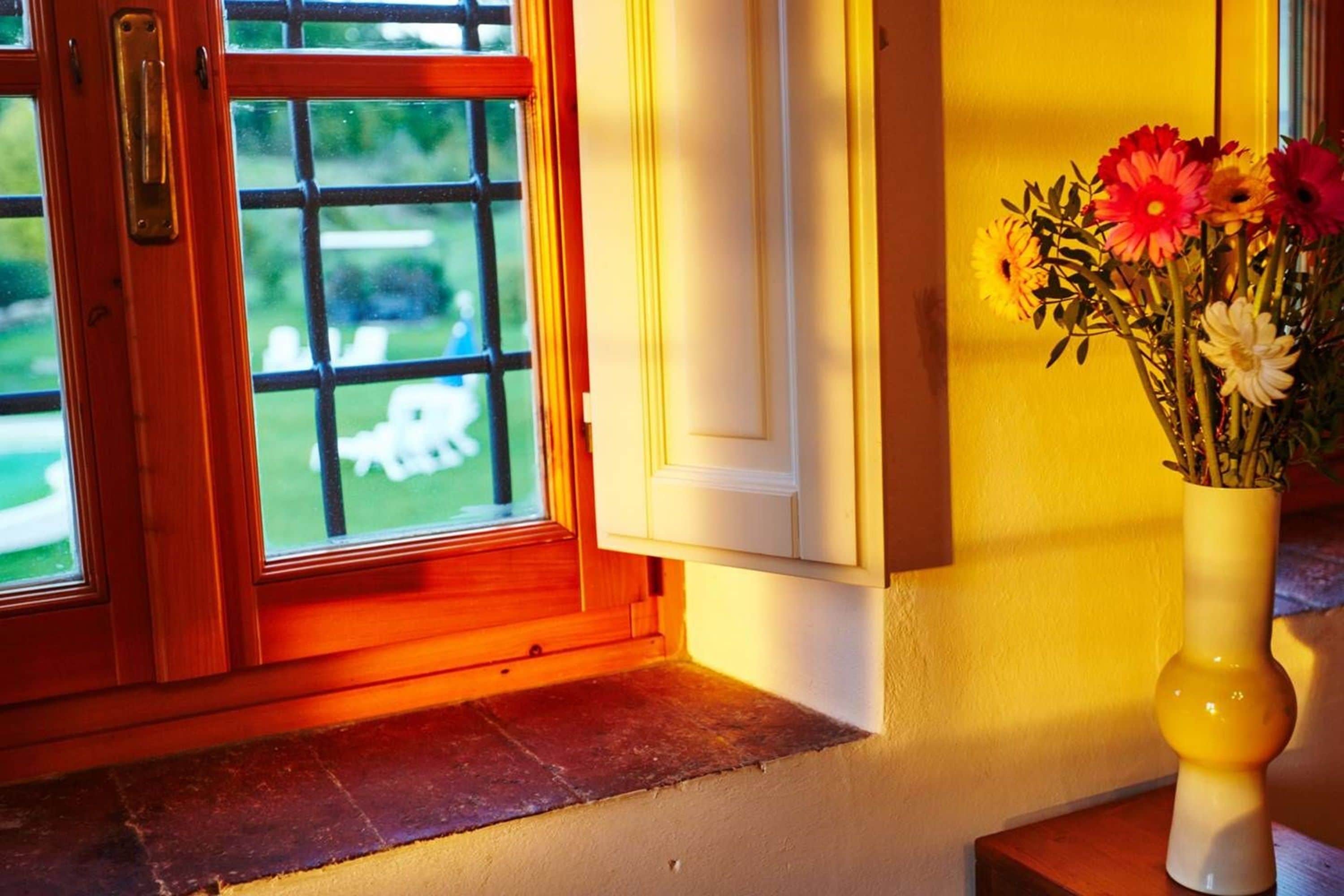 Una ventana con vistas a un césped y una silla blanca, un jarrón amarillo con flores sobre un alféizar de madera, y una puerta con pomo de latón.