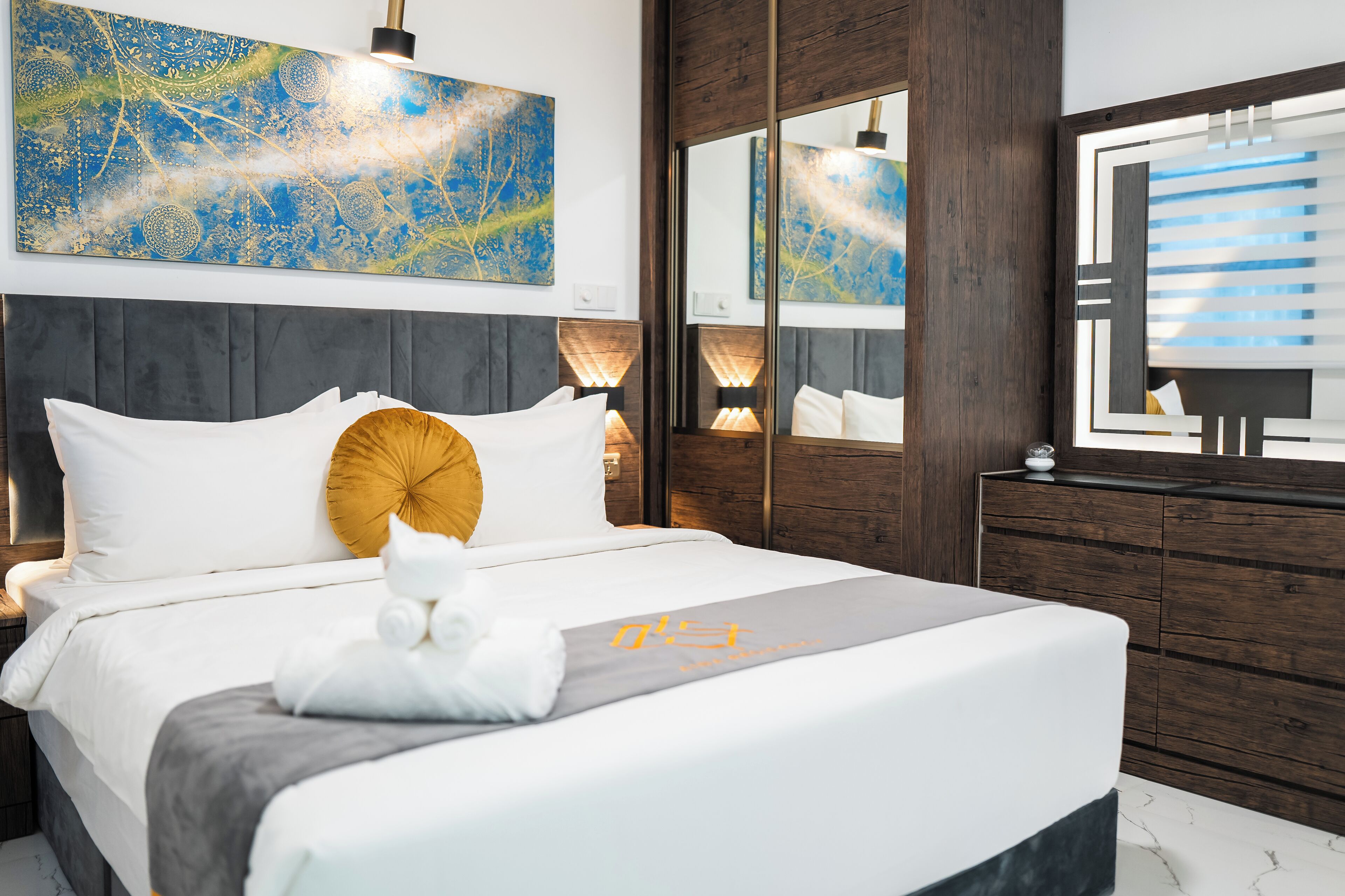 A modern hotel room with a large bed, a wooden headboard, a mirror, and a decorative wall piece.