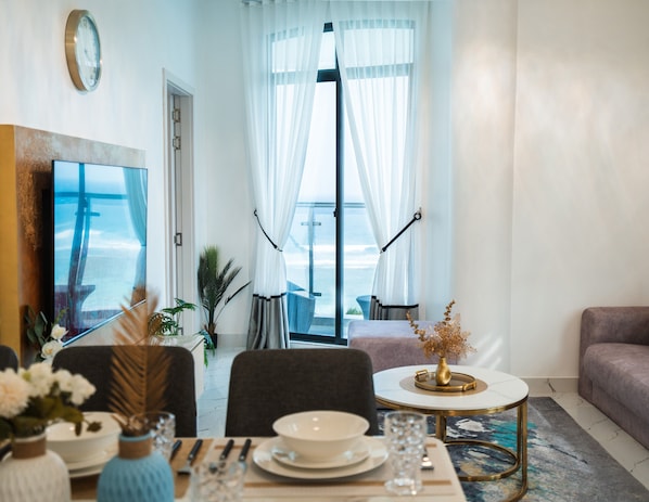 Ein modernes Wohnzimmer mit Essbereich, ein großes Fenster mit transparenten Vorhängen, Blick aufs Meer und ein runder Couchtisch mit einer Vase.