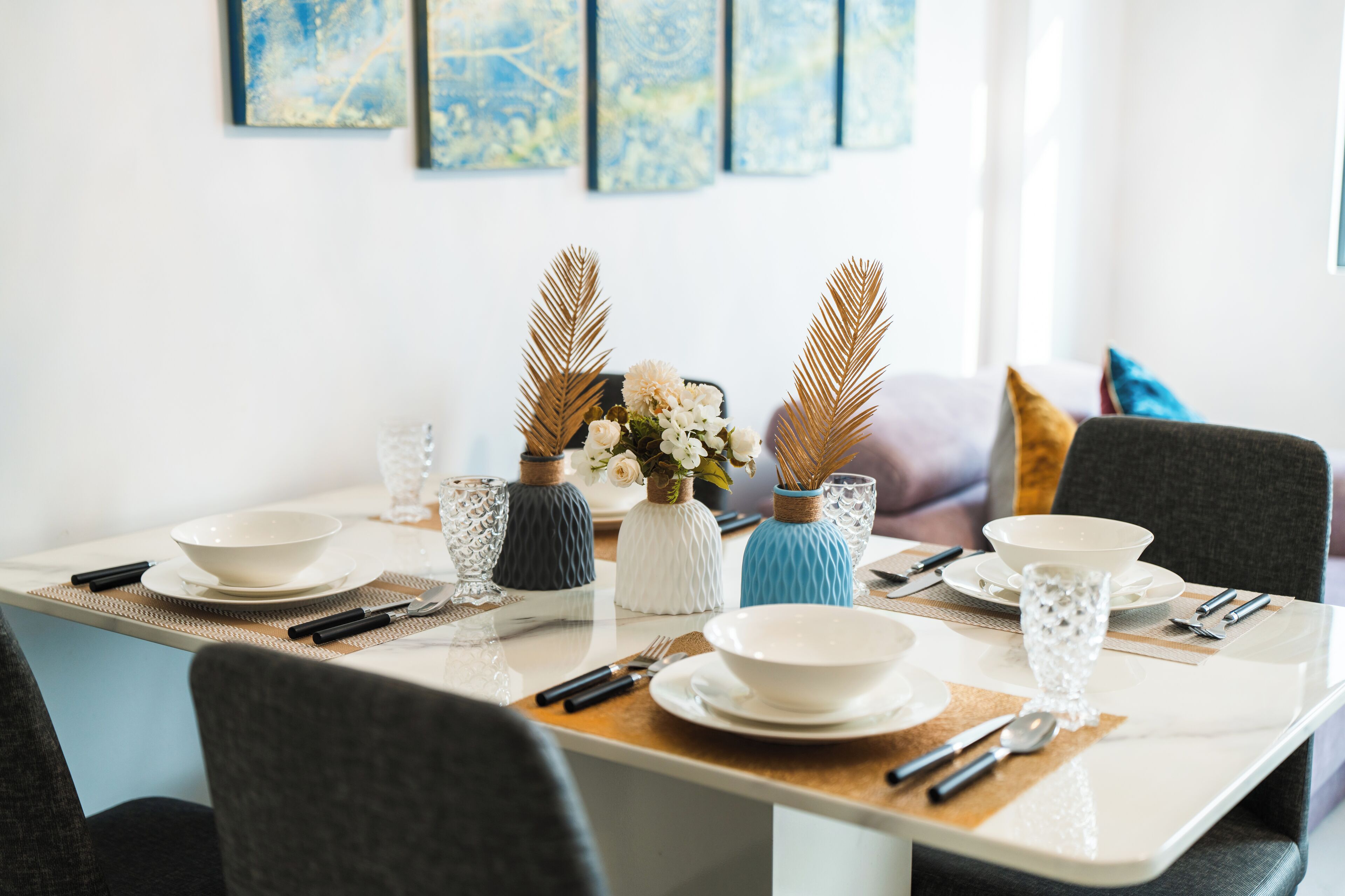 Une table à manger dressée avec de l’assiette, des bols et des couverts blancs, entourée de chaises.