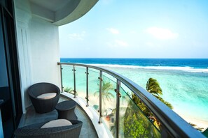 Een balkon met rieten stoelen en een glazen hekwerk met uitzicht op een strand en helder turquoise water, met zicht op de oceaan.