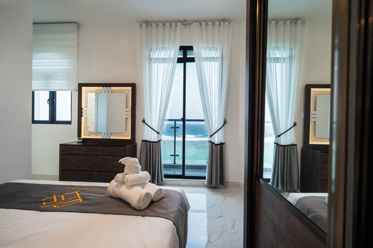 Habitación de hotel con cama, cómoda, espejo y vista al océano a través de una puerta de vidrio.