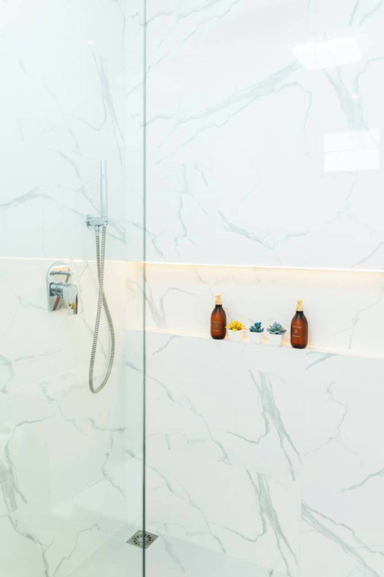 A glass-enclosed shower with a handheld showerhead and a wall-mounted faucet. There are two bottles on a shelf above the shower.