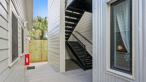 A narrow outdoor corridor with a wooden fence, a fire alarm box, and a staircase leading to an upper level.