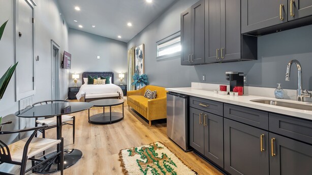 A modern hotel room with a bed, a yellow sofa, a dining table, and a kitchenette.