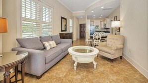 A living room with a gray sofa, a round coffee table, and a patterned armchair. There is a dining area in the background with a kitchen visible. - One bedroom with Oceanviews! (Saint Simons Island)