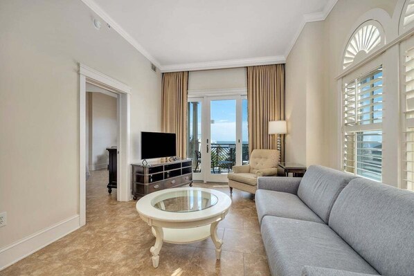A living room with a grey sofa, a glass coffee table, a flat-screen TV, and a balcony with curtains. - Sea Gate #308 One bedroom with Ocean views! (Saint Simons Island)