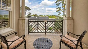 A balcony with two chairs and a table, overlooking a residential area with trees and a clear sky. - One Bedroom just across the street from the Beach! (Saint Simons Island)