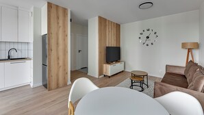 Une cuisine moderne avec des meubles blancs, un mur d’accent en bois, un téléviseur à écran plat et un coin repas comprenant une table ronde et des chaises.