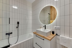 Un baño moderno con pared de azulejos blancos, tocador de madera con lavabo, espejo redondo y bañera.