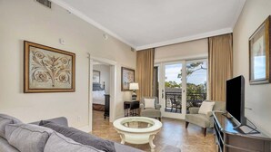 Ein modernes Wohnzimmer mit einem grauen Sofa, einem Couchtisch und einem Fernseher. Ein Balkon bietet Blick auf Bäume und ein Gebäude.