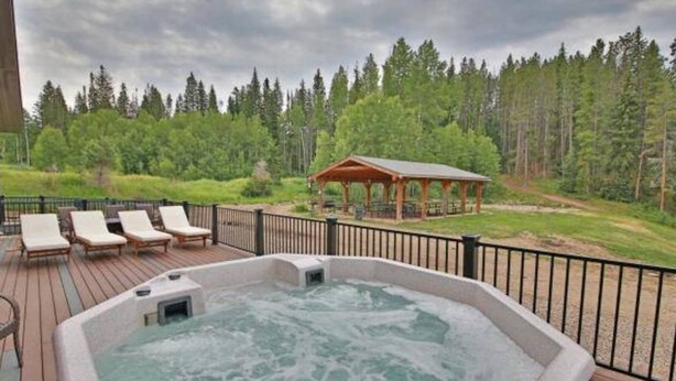 A hot tub on a wooden deck with lounge chairs, a gazebo, and a forested backdrop.