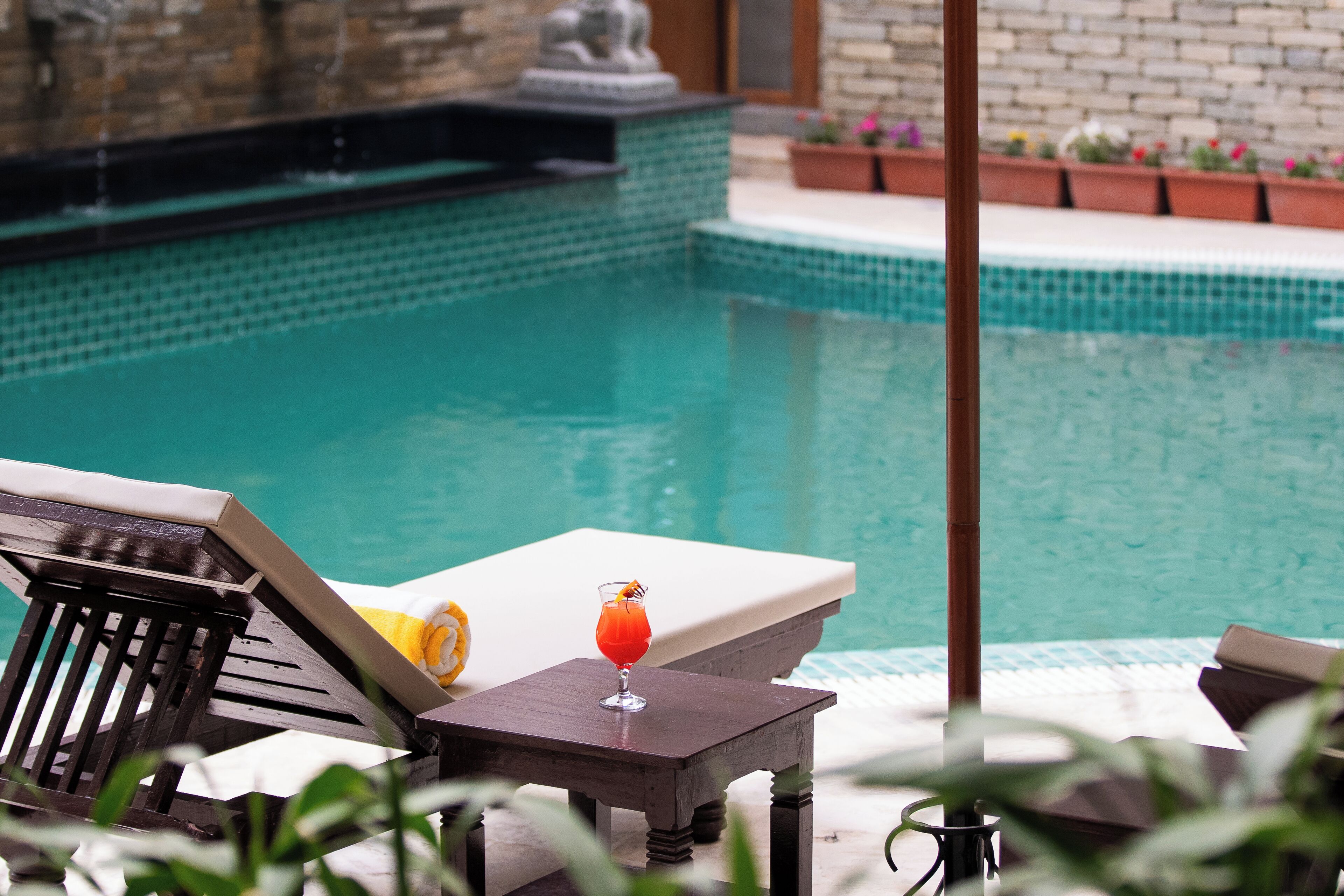 Zona de piscina con una tumbona, una mesa con una bebida y una estatua decorativa al fondo.