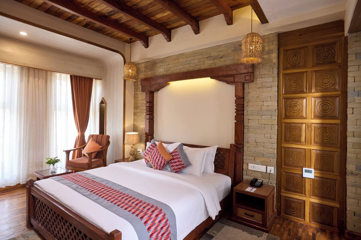 A hotel room with a large bed, wooden headboard, a brick accent wall, a wooden wardrobe, and a sitting area with a chair and table.