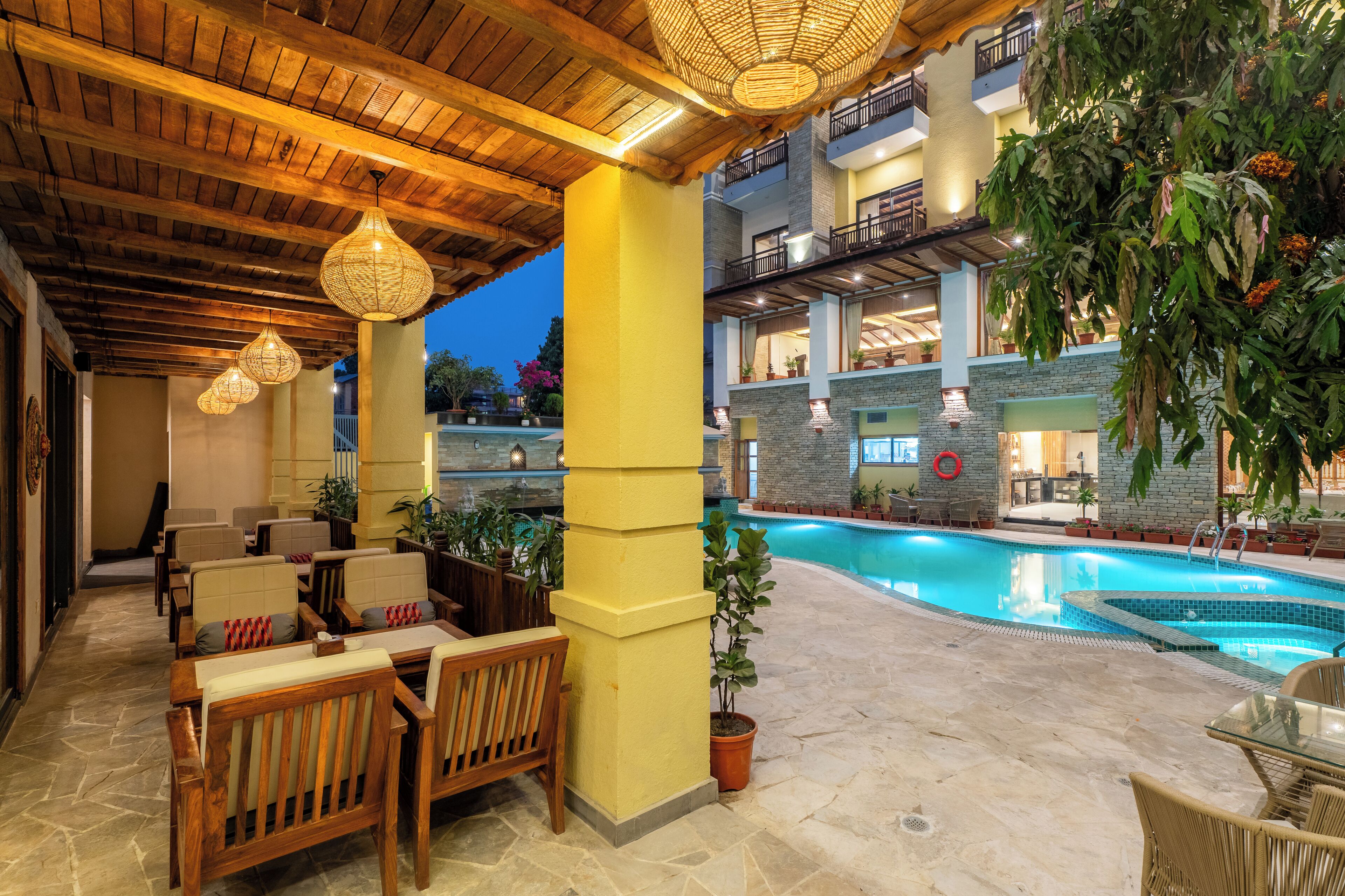 An indoor pool area with wooden furniture and a stone floor, surrounded by potted plants and a balcony overlooking the pool.