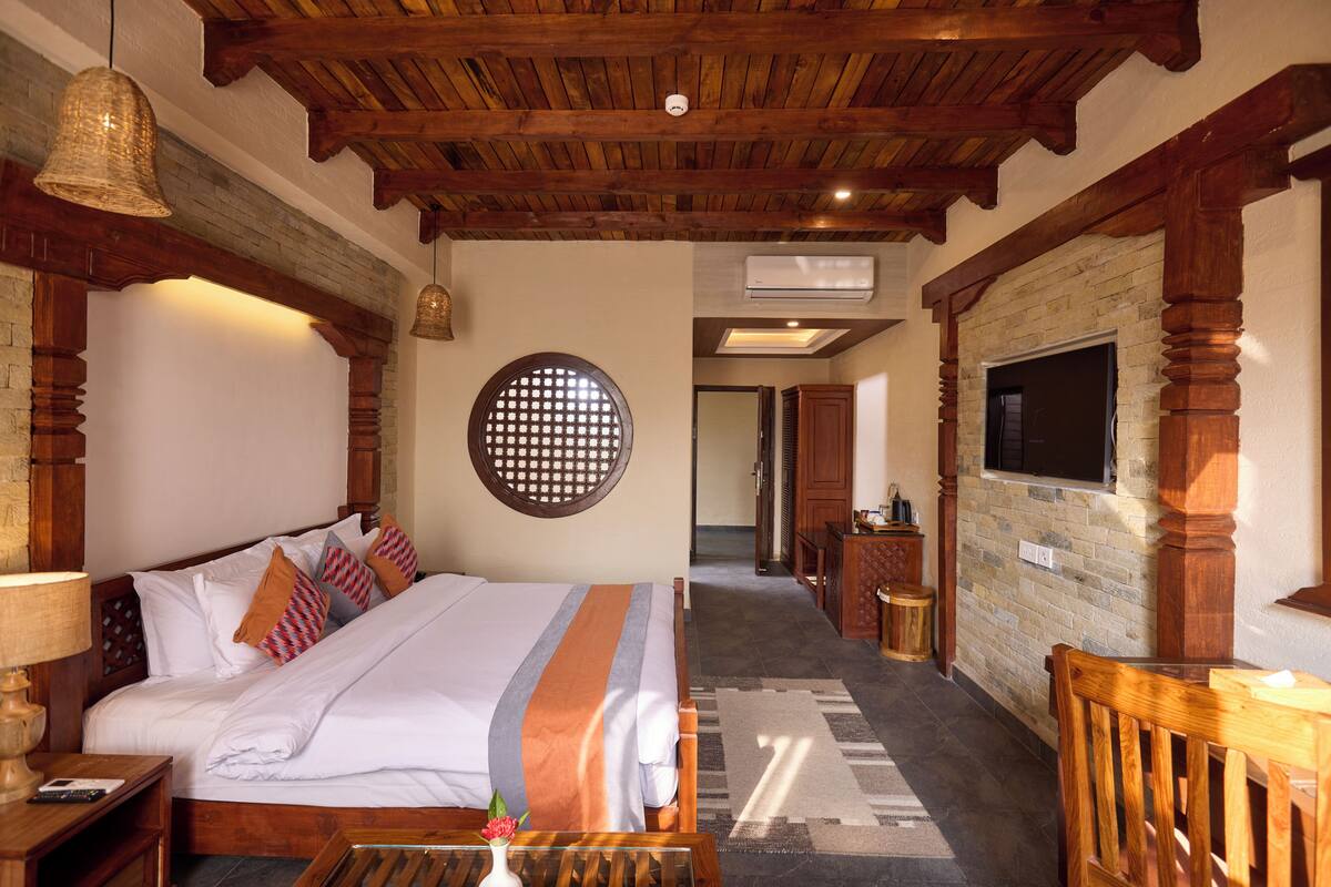 A hotel room with a wooden ceiling, a bed with white bedding, a round wall decoration, a TV mounted on a brick wall, and a wooden chair.