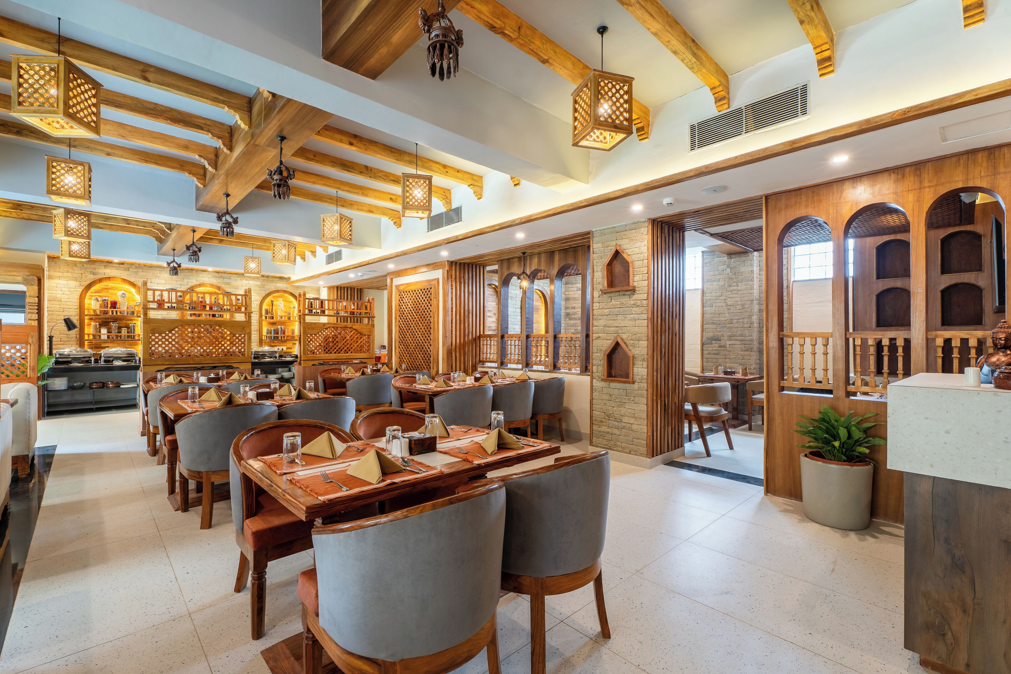 A restaurant with wooden beams, hanging lanterns, and a brick wall. Tables are set with chairs and centerpieces.