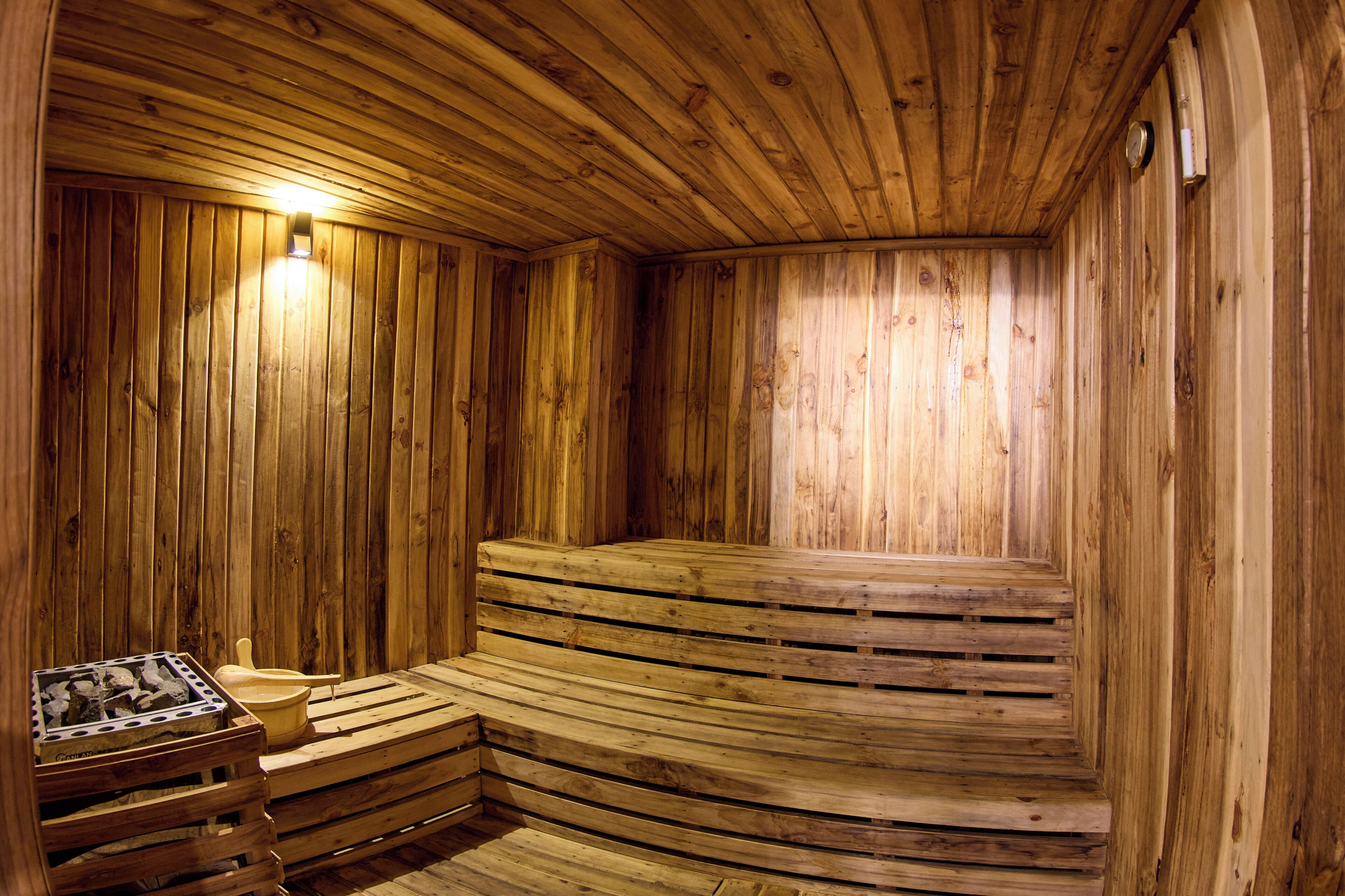 A wooden sauna with benches and a bench seat, a light fixture, and a wall-mounted heater.