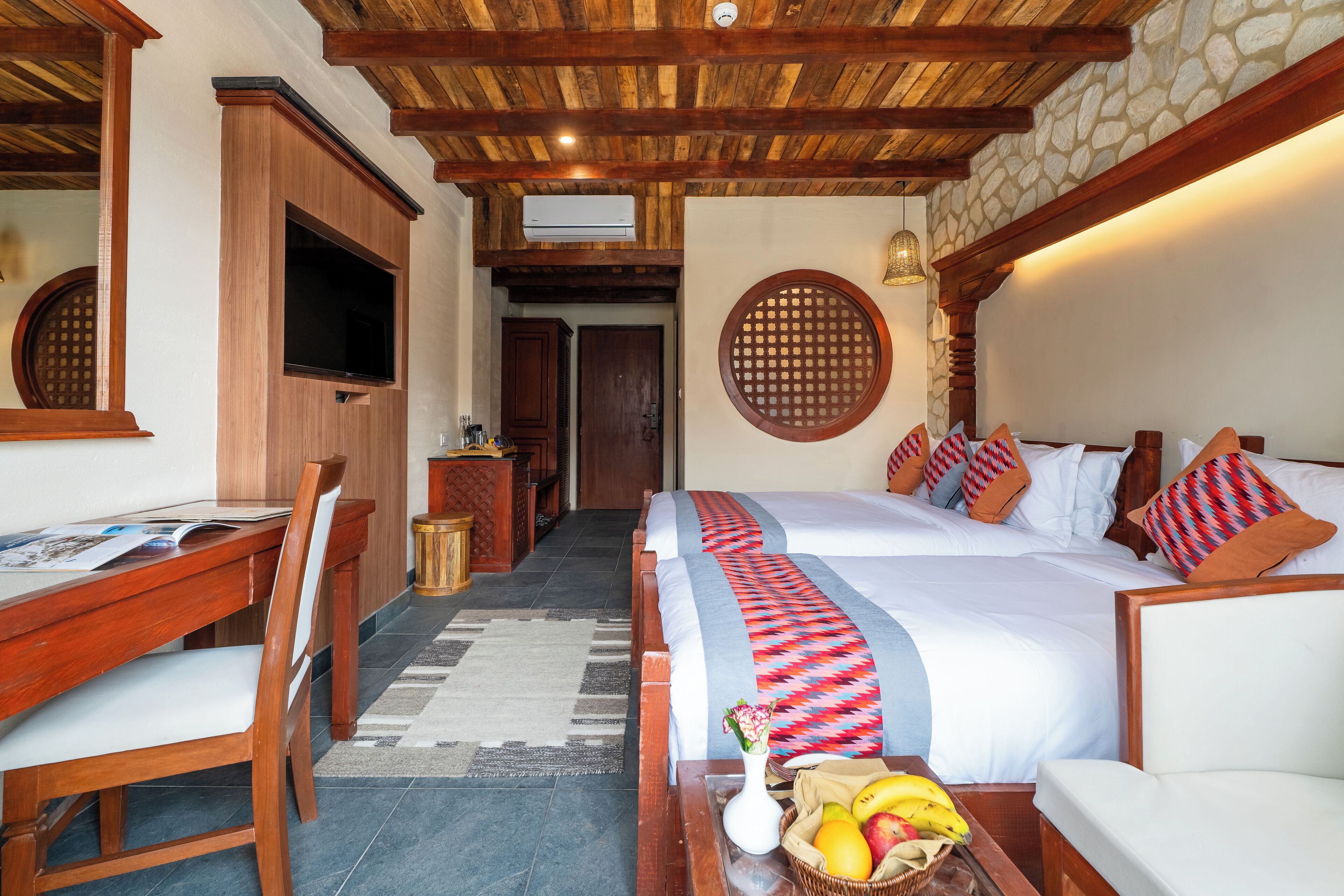 A hotel room with a wooden ceiling, a TV, a desk with a chair, a bed with patterned pillows, and a fruit basket on the nightstand.