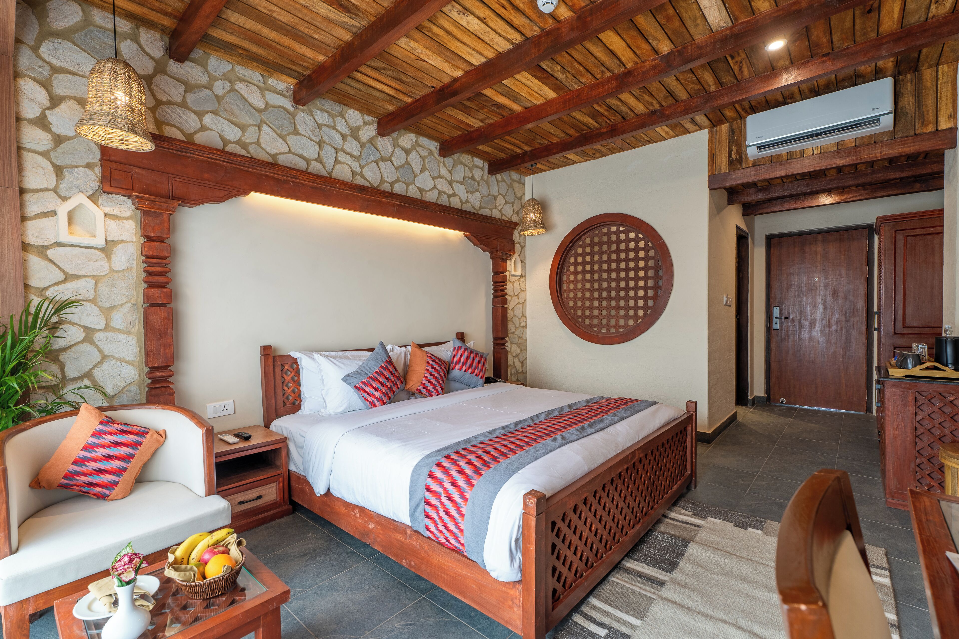 Un dormitorio con pared de piedra, vigas de madera en el techo, una cama con ropa de cama estampada, una mesita de noche, una cesta de mimbre con frutas y una pequeña mesa con un florero.