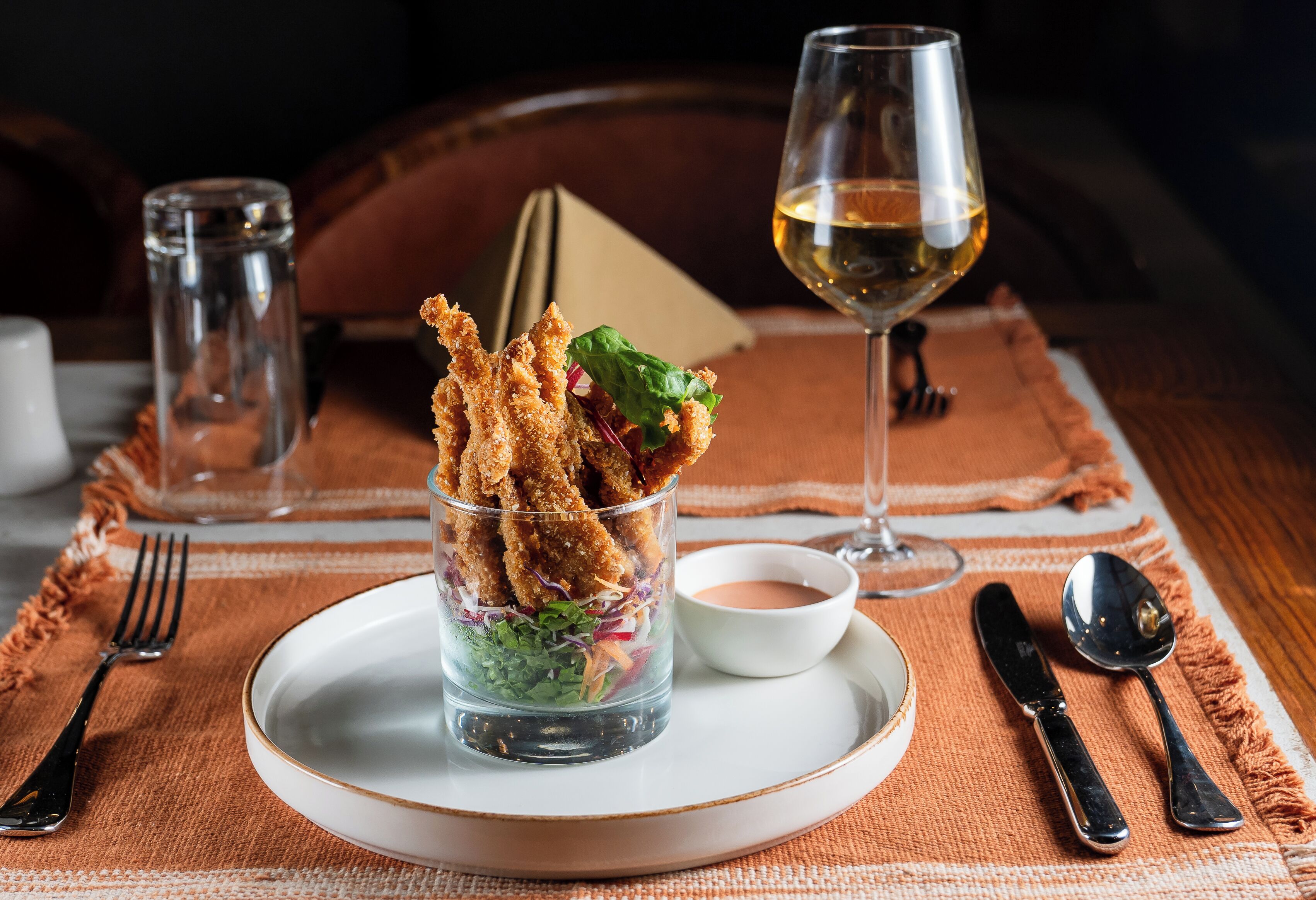 A glass of salad with breaded items and a small bowl of sauce on a plate, accompanied by a wine glass and cutlery on a table with a woven placemat.