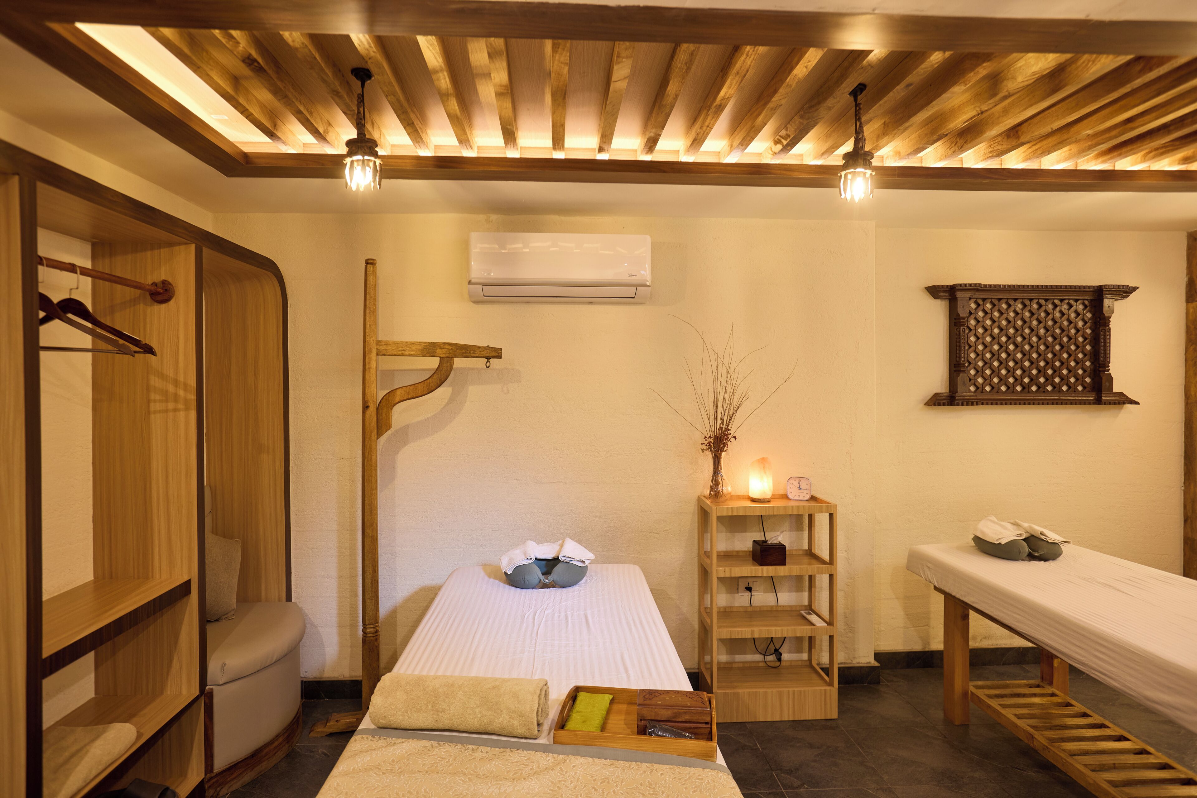 Una habitación con techo de madera, una cama con colchón blanco, un banco de madera, una estantería con un jarrón y un equipo de aire acondicionado.