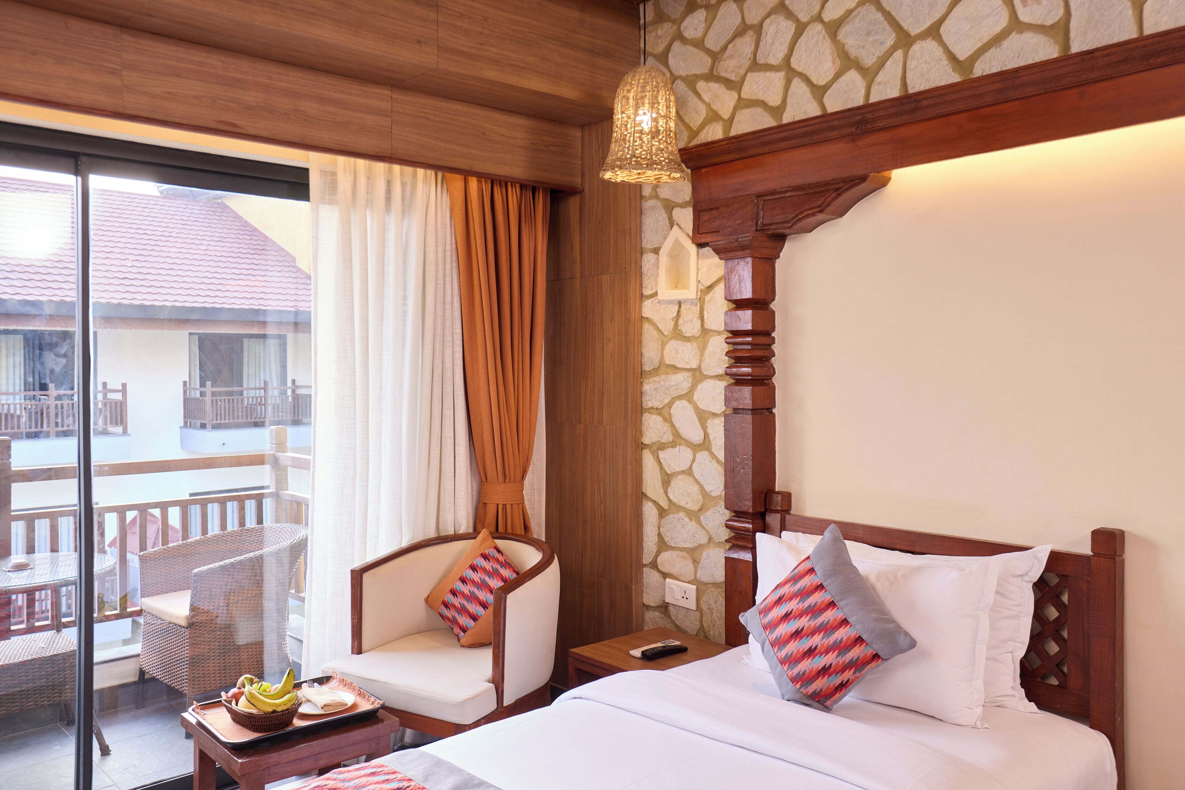 Quarto de hotel com cama, cadeira, mesa de cabeceira, bandeja com bananas e varanda com móveis de vime.