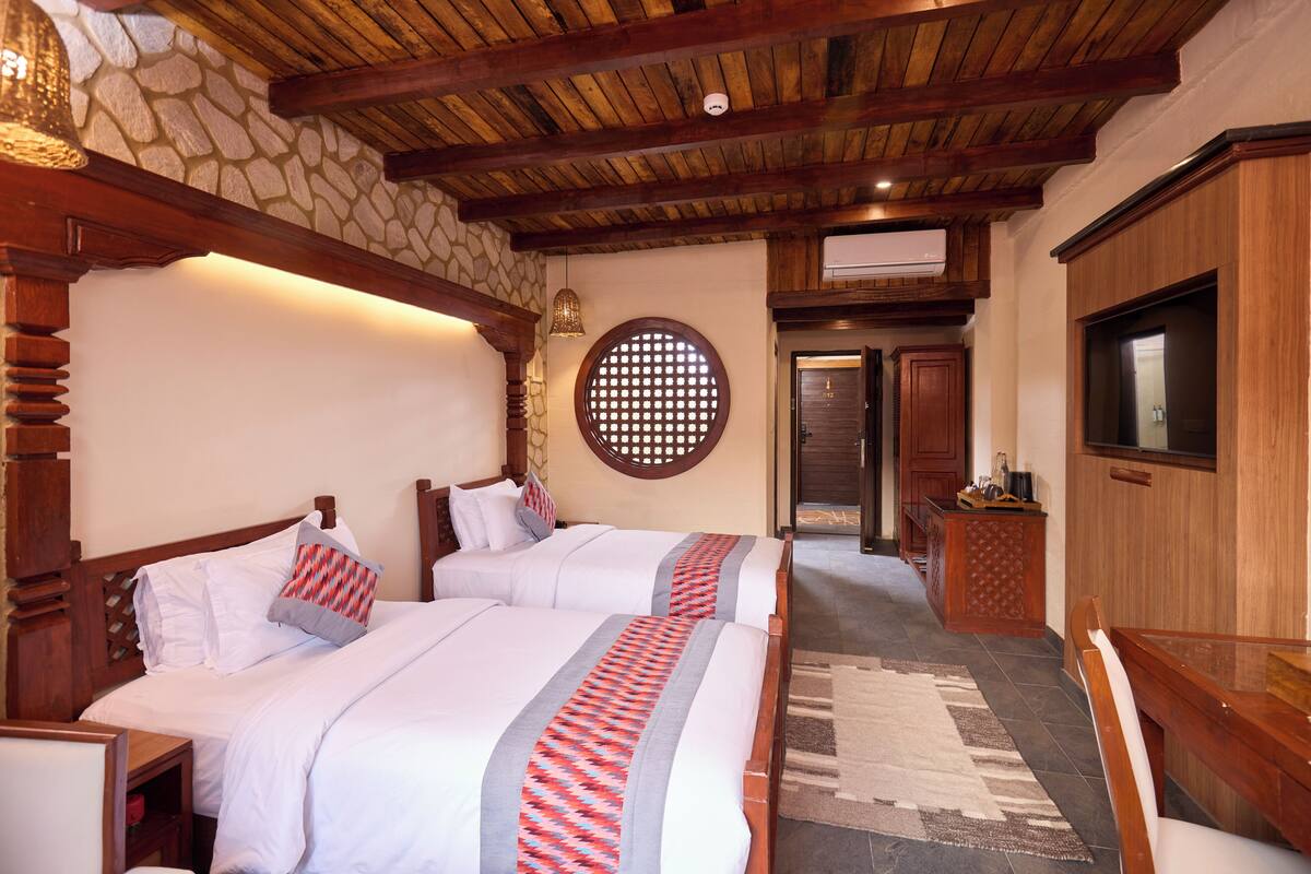 A hotel room with two beds, a stone wall, wooden ceiling beams, a round window, a TV, and a desk.
