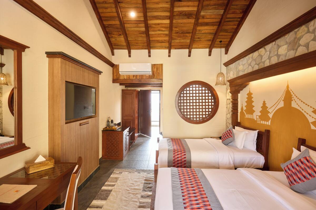 A hotel room with wooden beams, a large bed with patterned pillows, a desk with a mirror, and a stone wall with a decorative window.