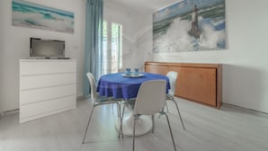 A dining area with a table set for two, a television on a cabinet, and a painting of a lighthouse on the wall.