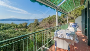 A balcony with a view of the sea and mountains, a table with a patterned tablecloth, and a green railing.