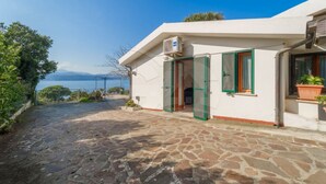 A stone-paved pathway leading to a white building with green shutters, a door, and an air conditioning unit. - APPARTAMENTI NEREIDI - MONO NEREIDI (Capoliveri)