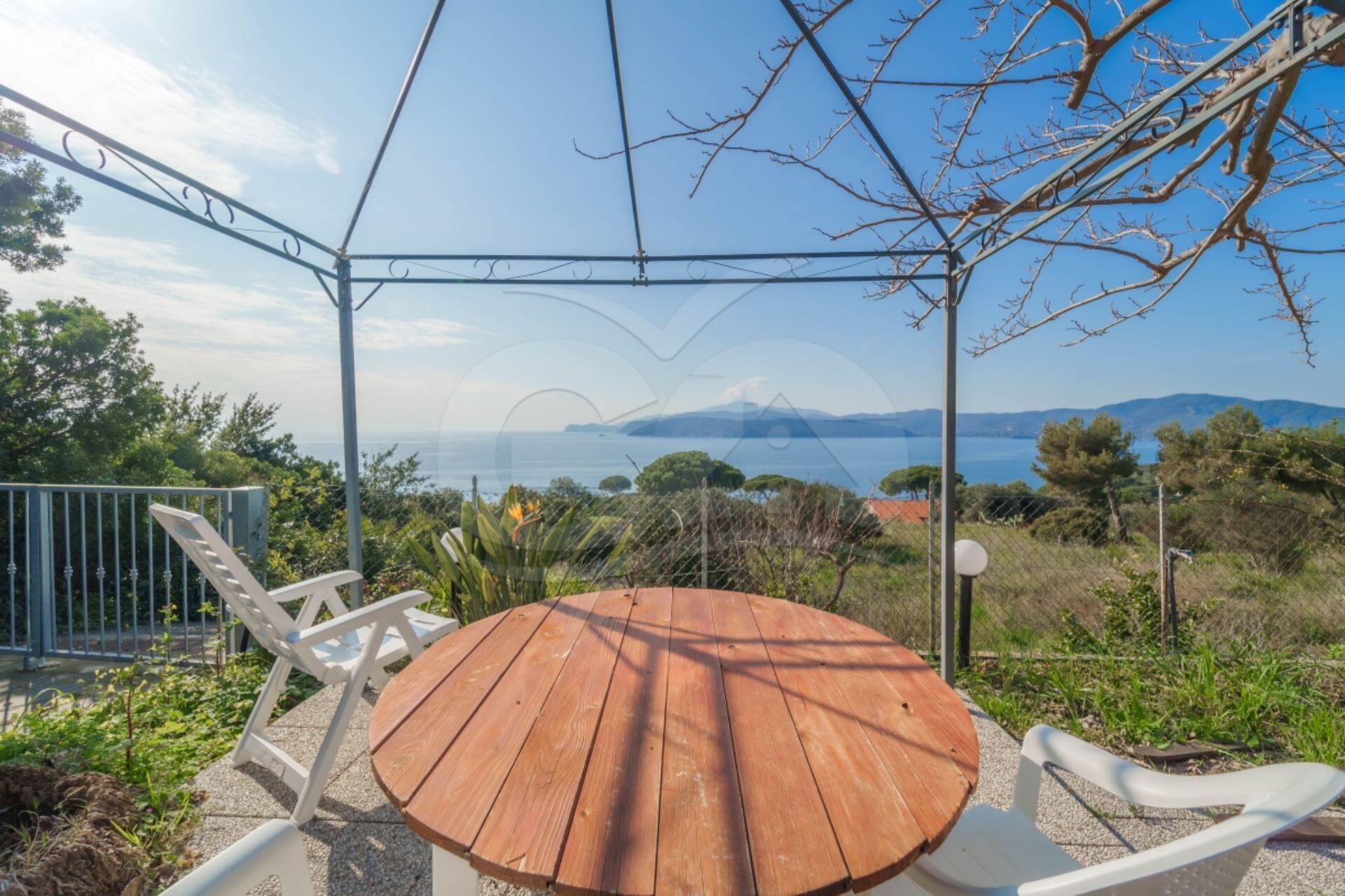 A wooden table and white chairs under a pergola with a view of the sea and mountains.