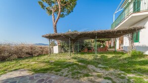 A stone patio leading to a covered outdoor area with a thatched roof, surrounded by greenery and a building with a balcony. - APPARTAMENTI NEREIDI - MONO NEREIDI (Capoliveri)