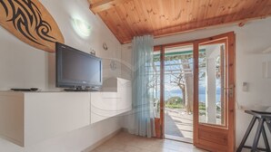 A room with a television on a white cabinet, a wooden ceiling, and a view of the outdoors through a glass door.