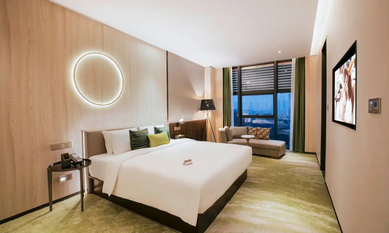A modern hotel room with a large bed, a seating area, a view of the city, and a circular light fixture on the wall.