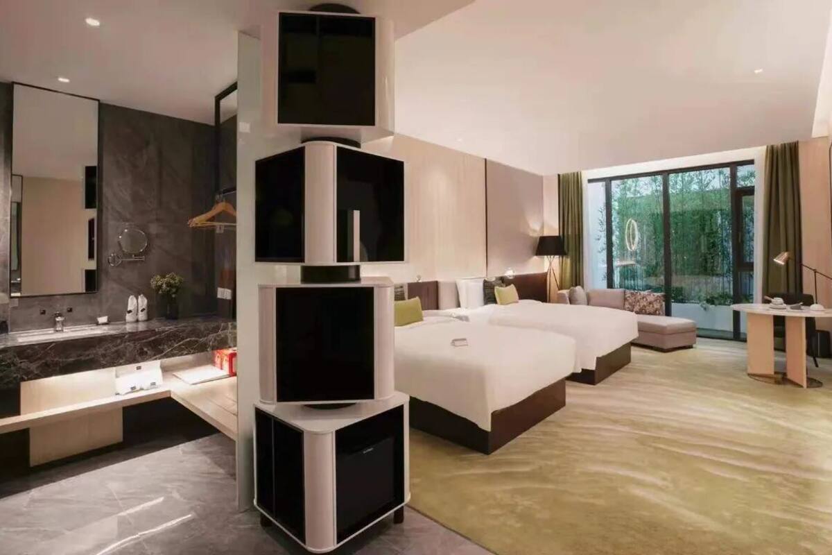 Moderni hotellihuone, jossa on suuri sänky, erillinen oleskelualue, peilillä varustettu meikkipöytä ja suuri ikkuna, josta on näkymä ulos.