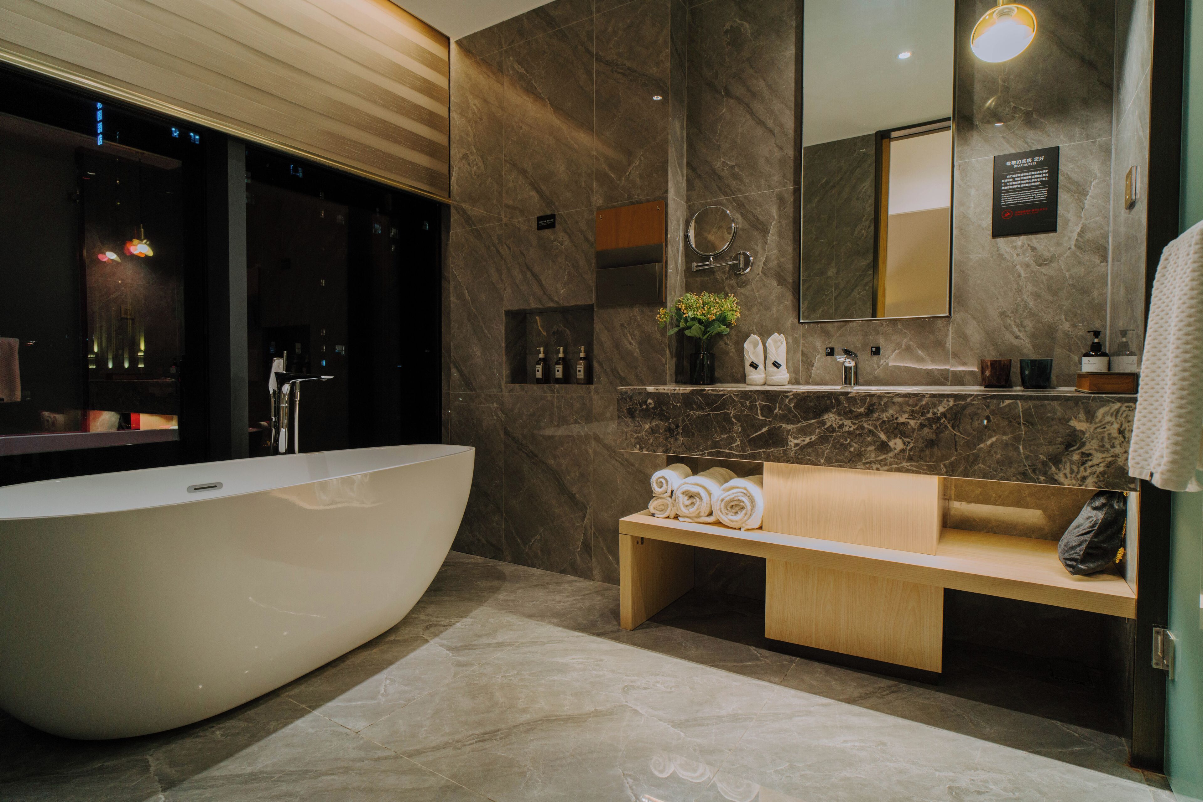 A modern bathroom with a freestanding bathtub, marble walls, a vanity with a mirror, and a bench with towels.