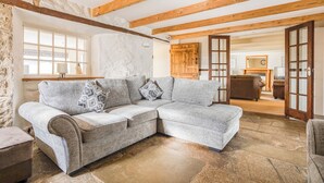 A cozy living room with a sectional sofa, stone flooring, exposed wooden beams, a fireplace, and a stone wall.