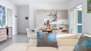 A modern kitchen with light wood cabinets, a dining area with a white table and chairs, and a beige sofa with decorative pillows.