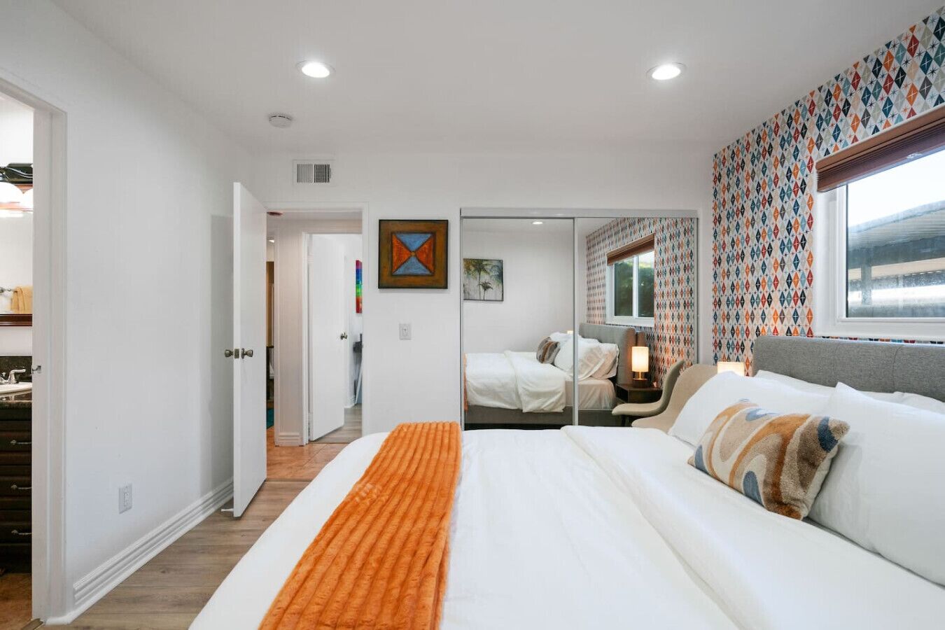 A modern bedroom with a large bed, a bedside table with a lamp, and a window with patterned curtains.