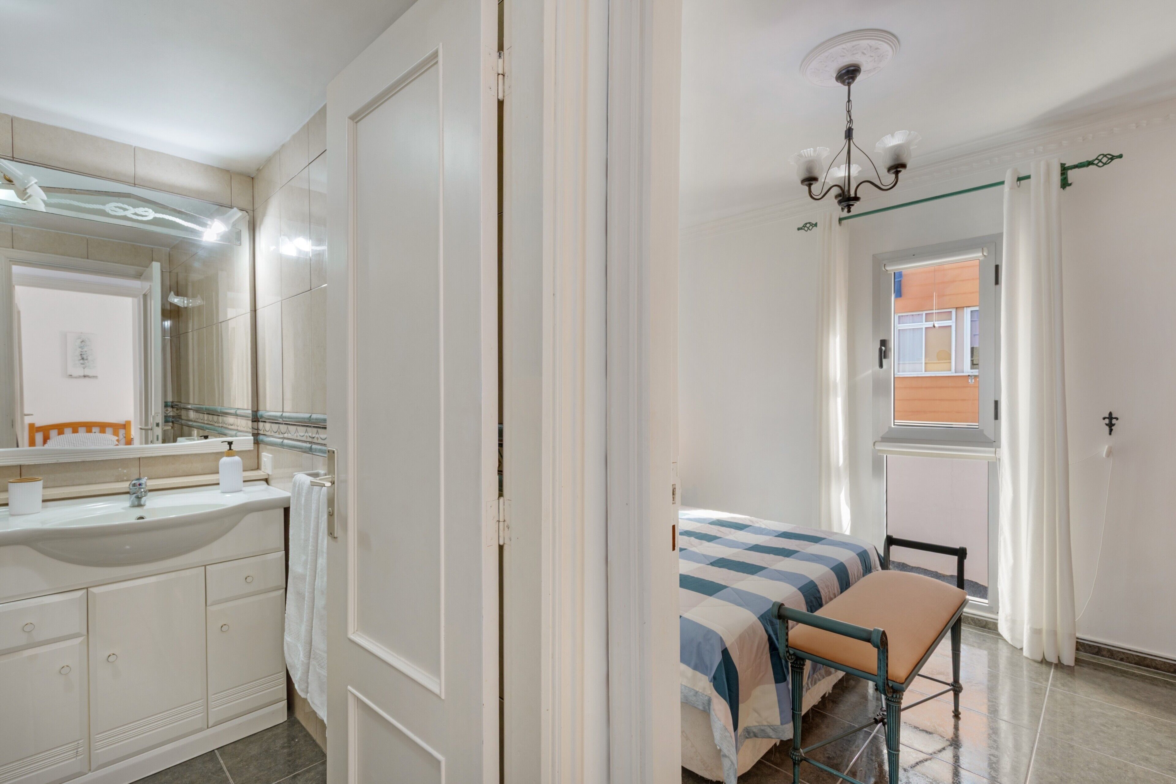 Un dormitorio con cama, ventana, baño con lavamanos y espejo, y puerta.