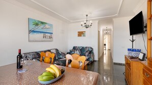 Un comedor con una mesa preparada para dos personas, un frutero y una botella de vino. Hay un sofá con un cuadro de una playa tropical.