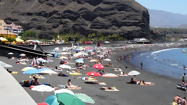 Una playa con gente tomando el sol y nadando, una colina rocosa al fondo y un cielo despejado.