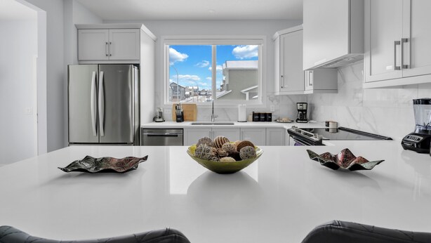 A modern kitchen with a large island, stainless steel appliances, and a view of the city through a large window. - Stylish 4-Bedroom & Fireplace (Mainfloor Bedroom & Full Bathroom) (Calgary)