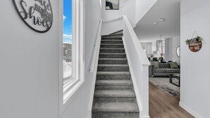 A modern interior with a staircase leading to a loft area, a living room with a sofa and coffee table, and a 'Please remove your shoes' sign on the wall. - Stylish 4-Bedroom & Fireplace (Mainfloor Bedroom & Full Bathroom) (Calgary)