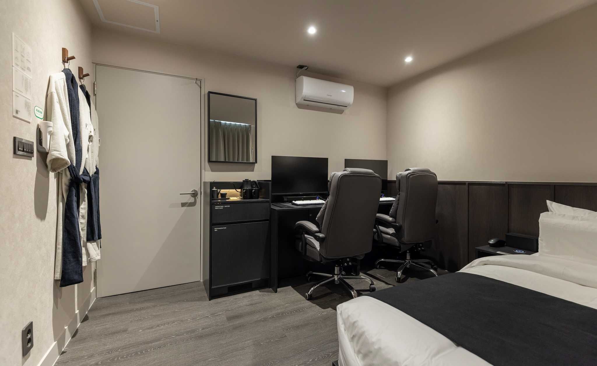 A hotel room with a bed, a desk with a computer, two chairs, a TV, and an air conditioning unit.