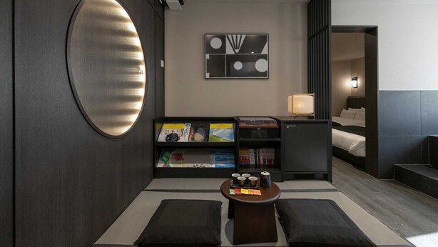 Una habitación de hotel moderna con un espejo redondo grande, una estantería con libros y revistas, una mesita con tazas y una cama con cabecero oscuro.